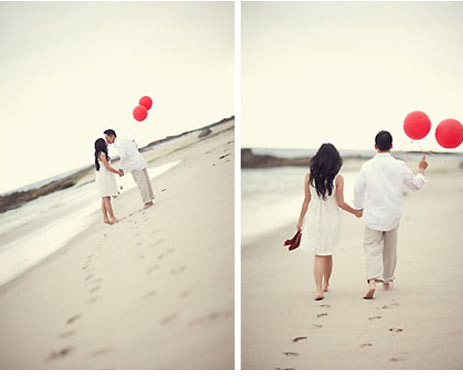 couple walking along beach with red balloons for engagement photo shoot 